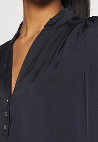 Mørkeblå bluse med v-udskæring, der har en rynket krave og lukning med fem knapper. Fremstillet af et glat, åndbart stof.