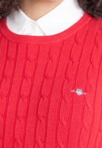 Gros plan d'une personne portant un pull rouge à torsades par-dessus une chemise blanche à col, avec un petit logo GANT brodé sur la poitrine.