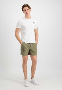 Junger Mann steht mit den Händen in den Taschen, trägt ein weißes T-Shirt, olivgrüne Cargo-Shorts und weiße Turnschuhe vor einem einfarbigen, hellen Hintergrund.
