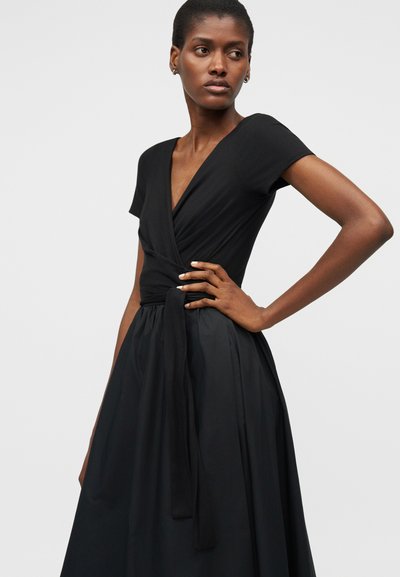 Γυναίκα με κοντά μαλλιά, ντυμένη με μαύρο φόρεμα τύπου wrap με κοντά μανίκια και δεμένη ζώνη στη μέση, στέκεται με το ένα χέρι στη μέση.