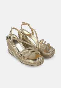Sandalias de cuña doradas con múltiples correas entrelazadas, base de cuerda texturizada y acabado metálico. Hebilla ajustable en el tobillo para un mejor ajuste.