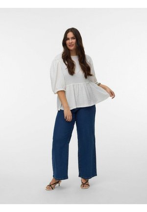 Kvinde med langt mørkt hår iført en hvid tekstureret bluse med pufærmer, blå vide jeans og sorte sandaler med remme, stående på hvid baggrund.