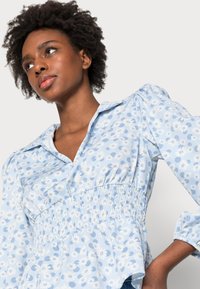 Blusa azul clara com um padrão floral, cintura franzida e mangas bufantes. Tecido suave com colarinho e fecho de botões na parte da frente.
