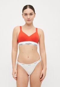 Červený bralet s širokým bílým elastickým páskem s nápisem "Calvin Klein" a odpovídající bílé tanga s podobným pásem s nápisem.