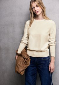 Maglione beige a maniche lunghe con colletto e polsini a coste, abbinato a jeans a gamba larga blu scuro. Il modello tiene una borsa marrone con texture.