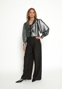 Metallisch zilveren cropped jas met opbollende mouwen en een striksluiting, gecombineerd met hoge zwarte broeken en puntige zwarte schoenen.
