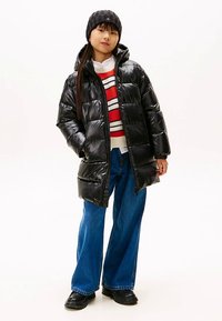 Giacca a sacco nera imbottita con zip, cappuccio e maniche lunghe. Indossata sopra un maglione a righe rosse e bianche e jeans larghi blu. Stivali neri.