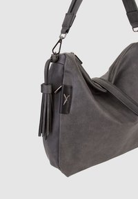 Sac à épaule gris texturé avec bandoulière réglable, fermeture zippée, détail de pompon sur le côté et petit logo métallique sur patch en cuir noir.