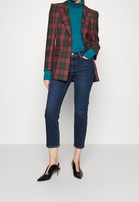 Blazer con estampado de cuadros rojo y verde, combinado con un cuello de tortuga color teja, jeans azul oscuro y tacones negros de punta. Diseño estructurado.