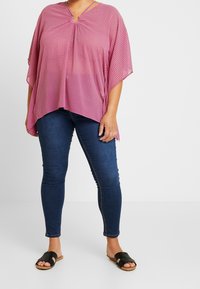 Haut ample à motif rose avec manches courtes, détail œillet au niveau de l'encolure, associé à un jean slim bleu foncé et des sandales noires à enfiler.