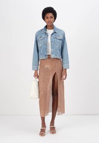 Denim jakna, bela majica, bleščeča rjava krila z razporkom do stegna in sandali z trakovi; drži belo torbico.