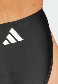 Crne kupaće gaćice od glatkog, rastezljivog materijala s visokim strukom. Sadrže bijeli Adidas logo u gornjem lijevom kutu.