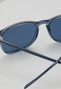 Ochelari de soare din plastic albastru, cu lentile întunecate, având un design clasic și brațe subțiri. Logo-ul Ray-Ban este vizibil pe braț.