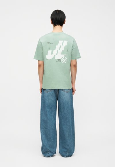 Άτομο με κοντά μαύρα μαλλιά που φοράει ένα μπλουζάκι μέντας με γραφικό και φαρδιά μπλε τζιν παντελόνια, στέκεται μπροστά σε ένα απλό λευκό φόντο.