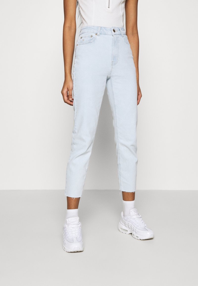 Vaqueros de tiro alto en azul claro con un corte ajustado y dobladillos deshilachados, combinados con zapatillas deportivas blancas y calcetines tobilleros. Sin patrones ni herrajes visibles.