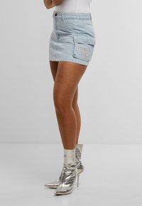 Hellblauer Jeansrock mit einer Seitentasche, verziert mit gesticktem "KANI." Kombiniert mit metallic-silbernen Knöchelstiefeln mit Stiletto-Absätzen.