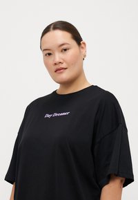 Camiseta negra de gran tamaño, hecha de algodón, con un gráfico de texto morado que dice "Day Dreamer" en el frente. Mangas cortas y cuello redondo.