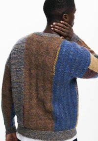 Pulover tricotat cu un design patchwork în nuanțe de cafea, albastru și bej, cu țesături texturate și cusături galbene contrastante pe marginile cusăturilor.