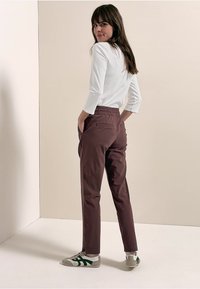 Mujer con cabello largo y castaño que lleva una camiseta blanca, pantalones marrones y zapatillas blancas con rayas verdes, de pie con las manos en los bolsillos, mirando hacia atrás.