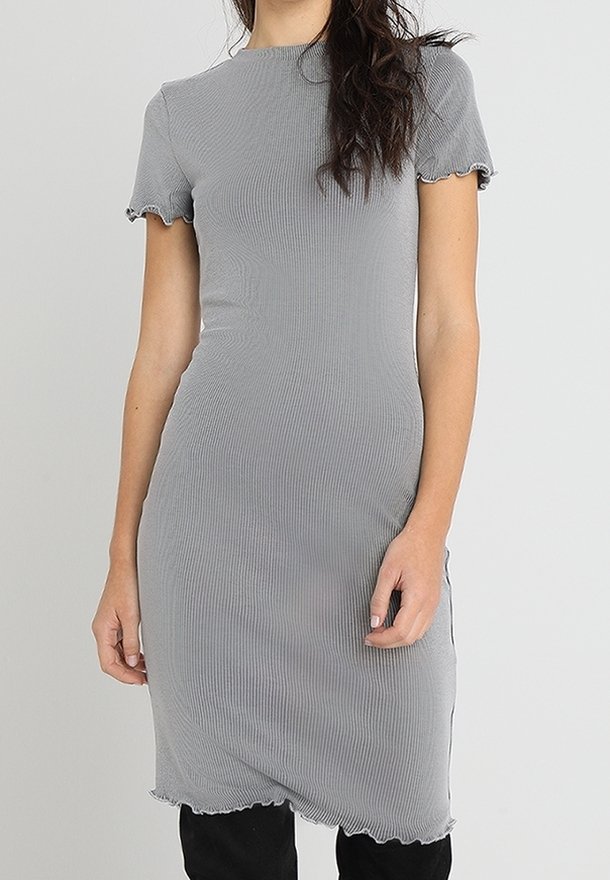 Femme portant une robe côtelée gris clair ajustée avec des manches courtes et des ourlets ondulés, debout devant un fond clair uni.