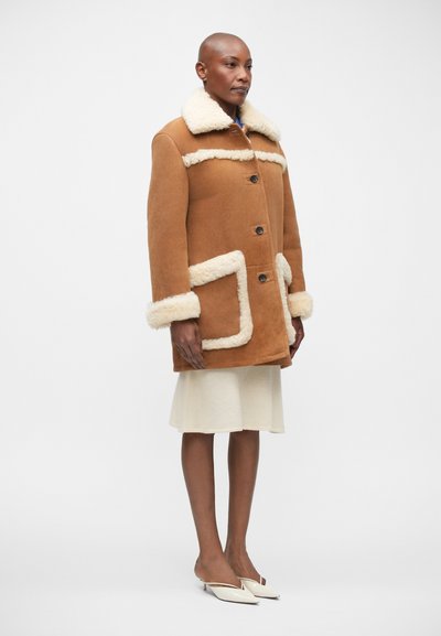 Cappotto in shearling marrone con colletto e bordi in pelliccia bianca, dotato di due tasche frontali, chiusura con bottoni e una vestibilità morbida. Indossato sopra una gonna leggera.