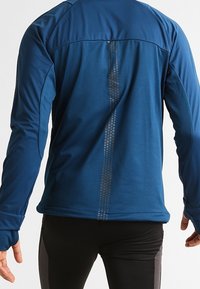 Uomo che indossa una giacca blu a maniche lunghe con fori per i pollici e una striscia riflettente verticale lungo la schiena, abbinata a pantaloni neri.