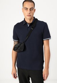 Mornarsko polo majica z belim proženim ovratnikom, kombinirana z majhno črno torbico čez ramo z verižnim ročajem in strukturirano površino.