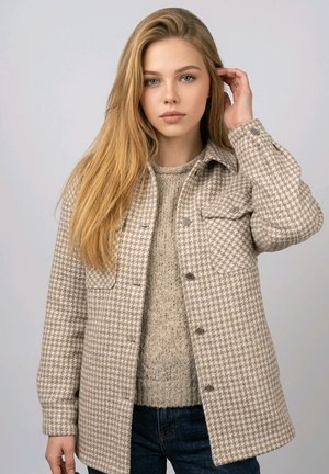 Joven con el pelo largo y rubio, lleva una chaqueta de pata de gallo beige y un suéter de punto, se ajusta el pelo con una mano, fondo neutro.