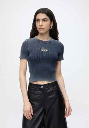 Jeune femme aux longs cheveux foncés portant un crop top bleu côtelé avec un petit détail métallique ovale, associé à un pantalon en cuir noir ample.