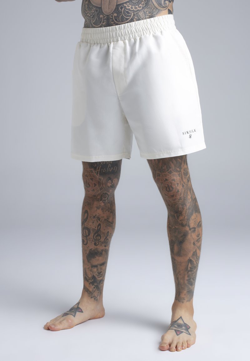 Gambe maschili con vari tatuaggi neri, indossano pantaloncini bianchi con elastico in vita e logo "SIKSILK" sulla coscia destra, a piedi nudi su sfondo semplice.