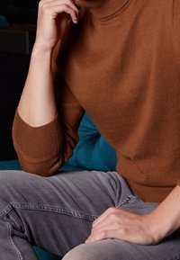 Brun strikket sweater med høj hals og ribbede manchetter, kombineret med grå jeans. Fokus på tekstur og farvekontrast.