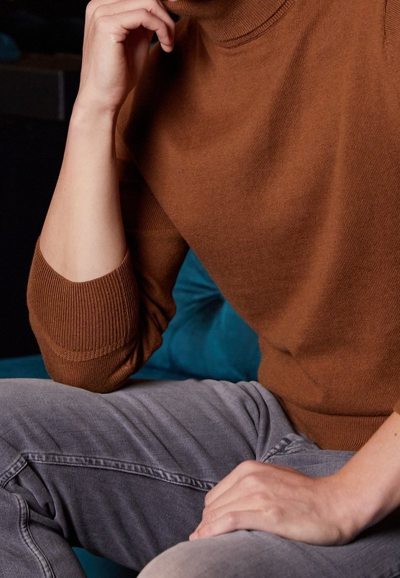 Brun strikket sweater med høj hals og ribbede manchetter, kombineret med grå jeans. Fokus på tekstur og farvekontrast.