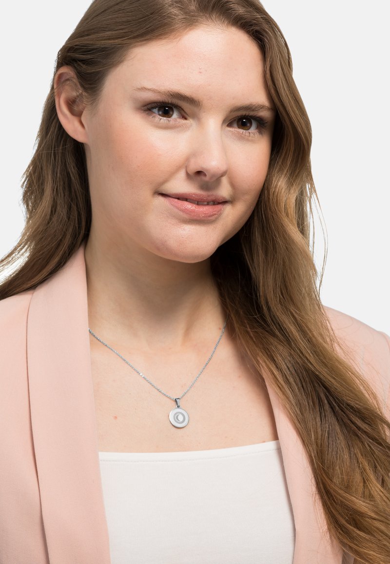 Srebrny naszyjnik z zawieszką w formie koncentrycznych kół na delikatnym łańcuszku, noszony na białej bluzce. Tło jest jasne jednolite.