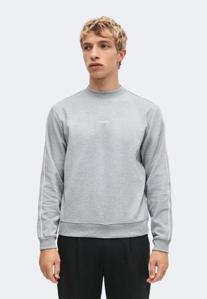 Młody mężczyzna z krótkimi blond włosami, noszący jasnoszary sweter Calvin Klein z białym obszyciem na rękawach oraz czarne spodnie.