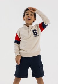 Felpa beige con zip, maniche rosse e blu, dotata di un logo e testo sul petto. Indossata con pantaloni cargo blu.