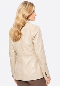 Beige leren jack met een aansluitend ontwerp, gladde textuur en knoopdetails op de mouwen. Gezien vanuit de achterkant.