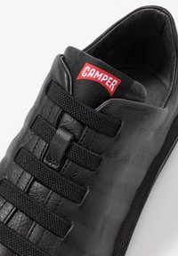 Black leather sneaker with a textured upper, elastic straps across the front, and a prominent red "CAMPER" tag on the tongue.