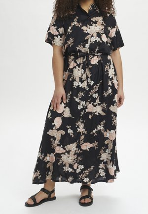 Kvinde iført en sort midi-kjole med blomsterprint, knapper og bindebånd i taljen og sorte sandaler, stående mod en ensfarvet hvid baggrund.