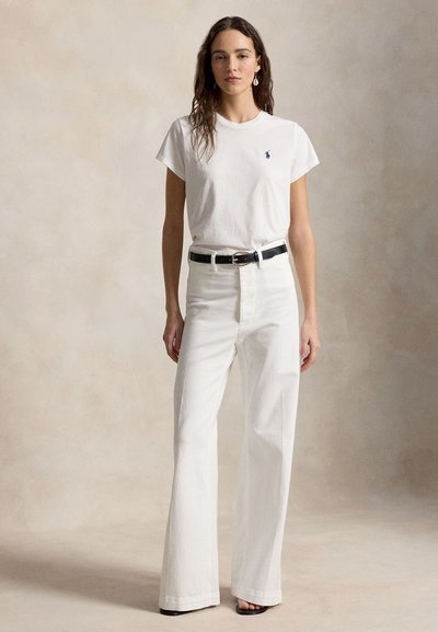 Γυναίκα που στέκεται φορώντας λευκό μπλουζάκι με μικρό λογότυπο, λευκό ψηλόμεσο παντελόνι με φαρδιά πόδια, μαύρη ζώνη και μαύρα παπούτσια με ανοιχτά δάχτυλα σε μπεζ φόντο.