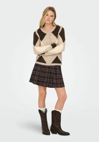 Cárdigan de punto beige y marrón con patrones geométricos, con botones en la parte delantera, combinado con una minifalda de cuadros negra y botas de vaquero marrones.