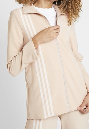 Nainen, joka on pukeutunut beigen väriseen vetoketjulliseen takkiin ja housuihin, joissa on valkoiset pystysuorat raidat, säätää takin vetoketjua.