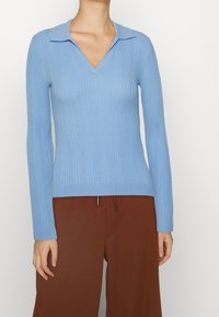 Pull en maille côtelée bleu clair avec col en V, doté de manches longues et d'une coupe ajustée, associé à un pantalon à jambes larges marron.