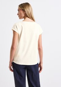 T-shirt crème à manches courtes avec un col rond et une coupe décontractée. Le tissu semble doux et présente une texture subtile.