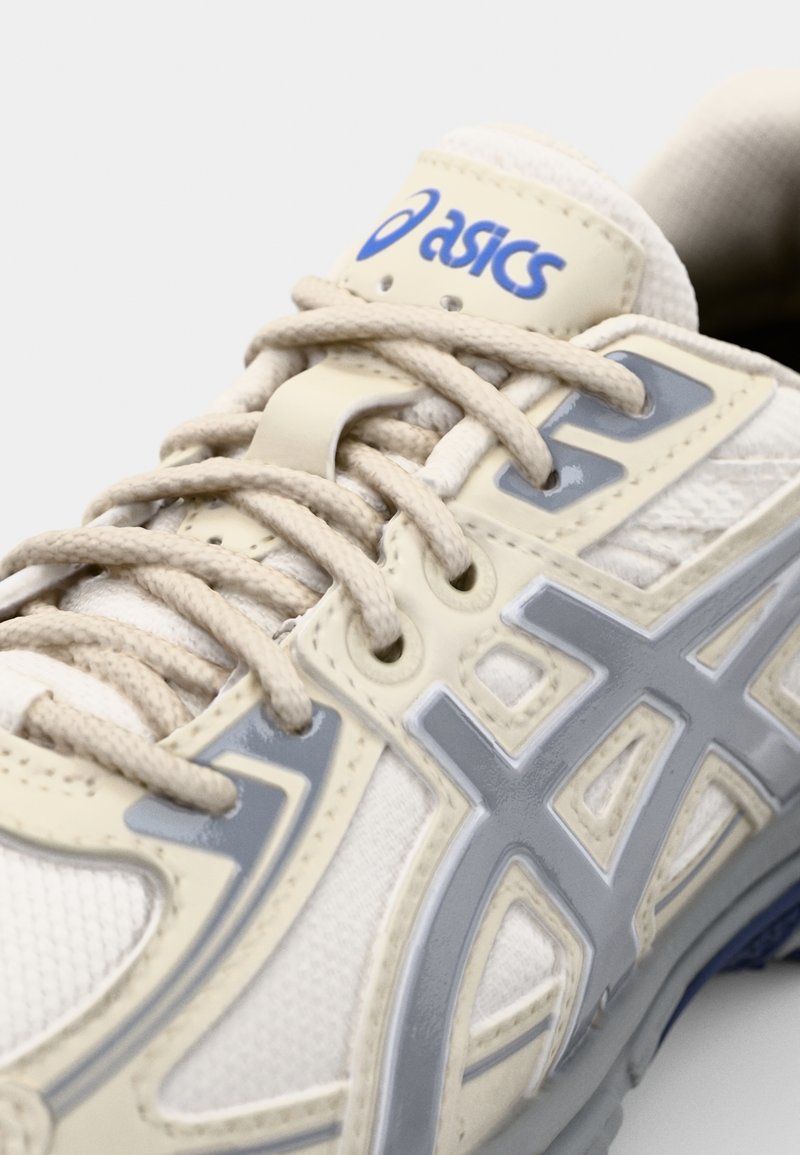 Zapatilla deportiva de color beige claro con detalles en gris, parte superior de malla texturizada, cordones planos y un logo de ASICS en la lengüeta. Suela de goma.