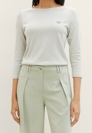 Vrouw draagt een lichtgrijs-wit gestreept shirt met lange mouwen en een klein zwart hartje op de borst, gecombineerd met lichtgroene broek met hoge taille.