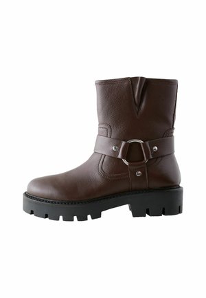 Bottine en cuir marron avec semelle noire épaisse et bande décorative avec détail anneau en métal sur le côté.