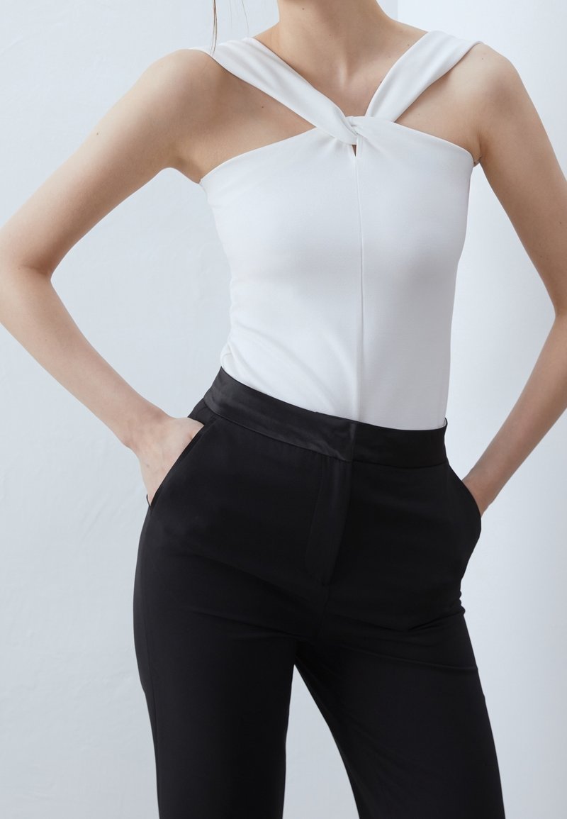Femme portant un haut blanc sans manches avec des bretelles torsadées et un pantalon noir taille haute, les mains dans les poches, debout devant un fond uni.