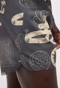 Σκούρα υφασμάτινα σορτς με αφηρημένα μπεζ σχήματα και σχέδια που φοριούνται από ένα άτομο, εμφανίζοντας το κάτω μέρος του μηρού και λεπτομέρειες της πλαϊνής ραφής.