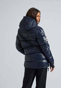 Marinsblå puffer-jacka med avtagbar huva, quiltat mönster, glänsande ytfinish, sidoventiler och logopatch på vänster ärm.