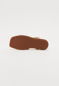 Sandalmit's såle har en strukturert brun gummibunn. Den har en glatt beige overdel og et detaljert dekorativt element langs siden.